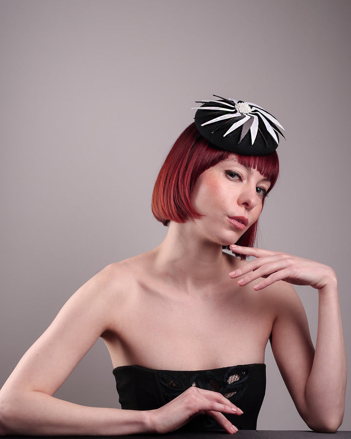 The model showcases the front of the Corvina pillbox fascinator, which is designed in a classic black button-shaped felt, featuring a dynamic swirl of sharp white and black feather blades that radiate from a white beaded centre. Designed by Merve Bayindir.