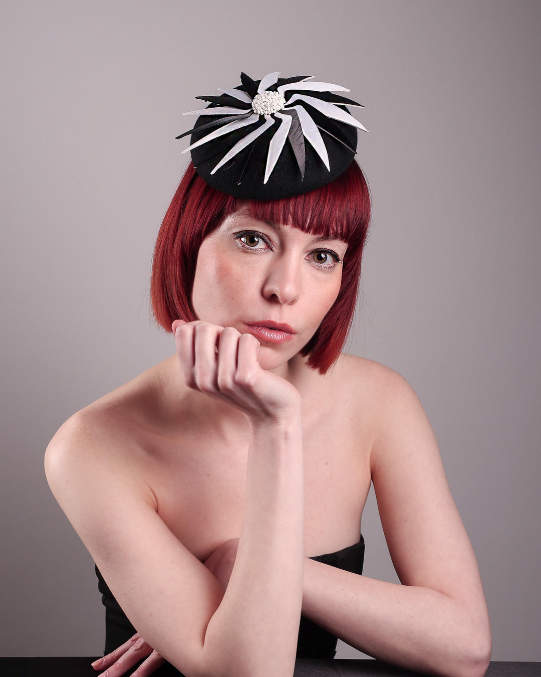 The model showcases the front straight of the Corvina pillbox fascinator, which is designed in a classic black button-shaped felt, featuring a dynamic swirl of sharp white and black feather blades that radiate from a white beaded centre. Designed by Merve Bayindir.