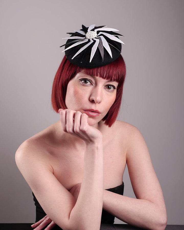The model showcases the front straight of the Corvina pillbox fascinator, which is designed in a classic black button-shaped felt, featuring a dynamic swirl of sharp white and black feather blades that radiate from a white beaded centre. Designed by Merve Bayindir.