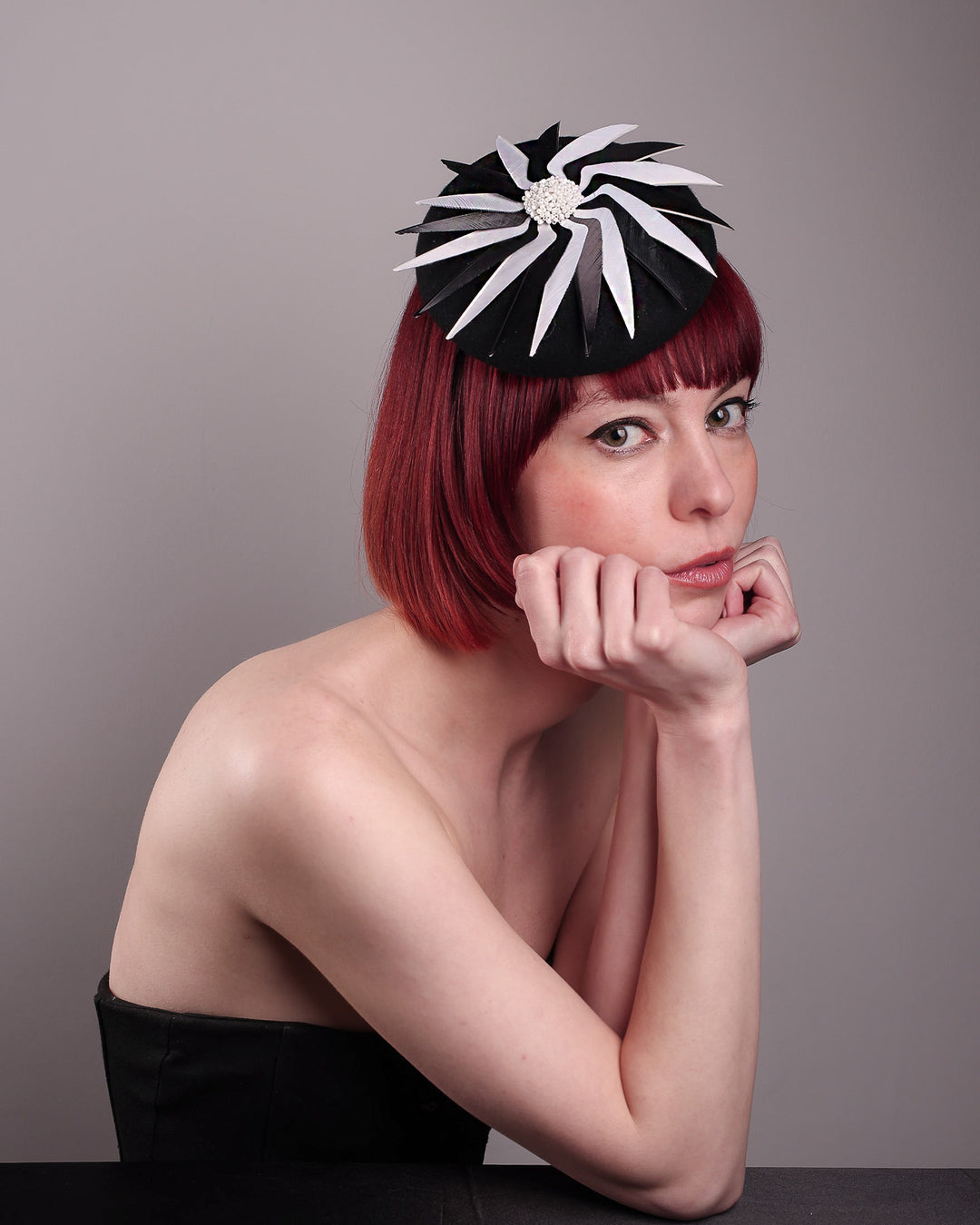 The model showcases the right front of the Corvina pillbox fascinator, which is designed in a classic black button-shaped felt, featuring a dynamic swirl of sharp white and black feather blades that radiate from a white beaded centre. Designed by Merve Bayindir.