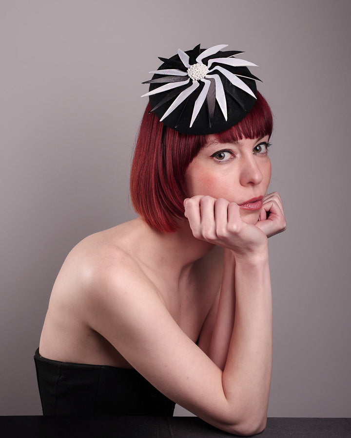 The model showcases the right front of the Corvina pillbox fascinator, which is designed in a classic black button-shaped felt, featuring a dynamic swirl of sharp white and black feather blades that radiate from a white beaded centre. Designed by Merve Bayindir.
