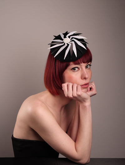 The model showcases the right front of the Corvina pillbox fascinator, which is designed in a classic black button-shaped felt, featuring a dynamic swirl of sharp white and black feather blades that radiate from a white beaded centre. Designed by Merve Bayindir.