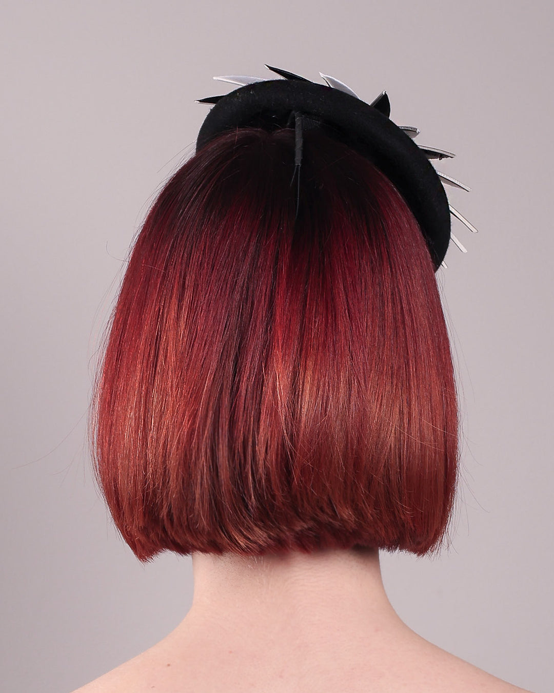The model showcases the back of the Corvina pillbox fascinator, which is designed in a classic black button-shaped felt, featuring a dynamic swirl of sharp white and black feather blades that radiate from a white beaded centre. Designed by Merve Bayindir.