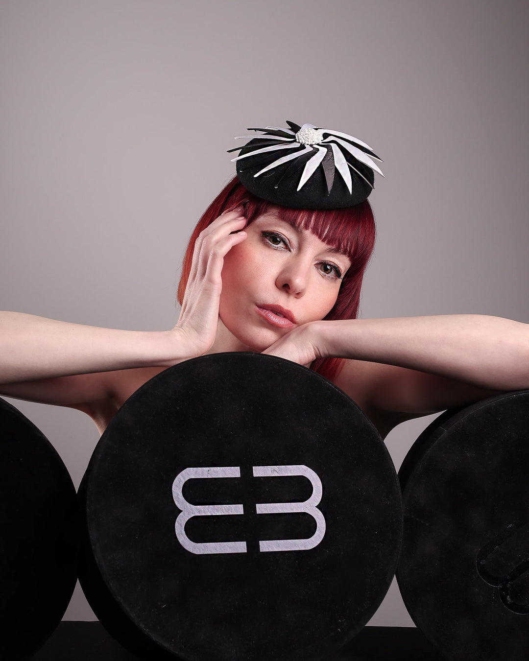 The model poses with Merve Bayindir hat boxes while showcasing the front of the Corvina pillbox fascinator, which is designed in a classic black button-shaped felt, featuring a dynamic swirl of sharp white and black feather blades that radiate from a white beaded centre. Designed by Merve Bayindir.