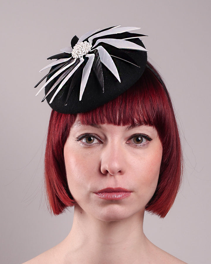 The model showcases the right front close up of the Corvina pillbox fascinator, which is designed in a classic black button-shaped felt, featuring a dynamic swirl of sharp white and black feather blades that radiate from a white beaded centre. Designed by Merve Bayindir.
