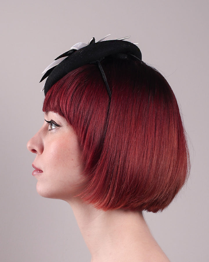 The model showcases the left side close up of the Corvina pillbox fascinator, which is designed in a classic black button-shaped felt, featuring a dynamic swirl of sharp white and black feather blades that radiate from a white beaded centre. Designed by Merve Bayindir.
