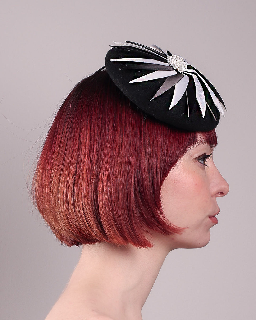 The model showcases the right side close up of the Corvina pillbox fascinator, which is designed in a classic black button-shaped felt, featuring a dynamic swirl of sharp white and black feather blades that radiate from a white beaded centre. Designed by Merve Bayindir.