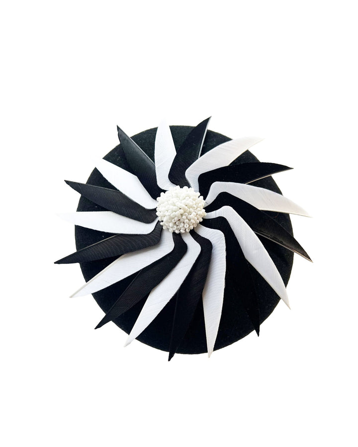 Still life picture of the Corvina pillbox fascinator, which is designed in a classic black button-shaped felt, featuring a dynamic swirl of sharp white and black feather blades that radiate from a white beaded centre. Designed by Merve Bayindir.