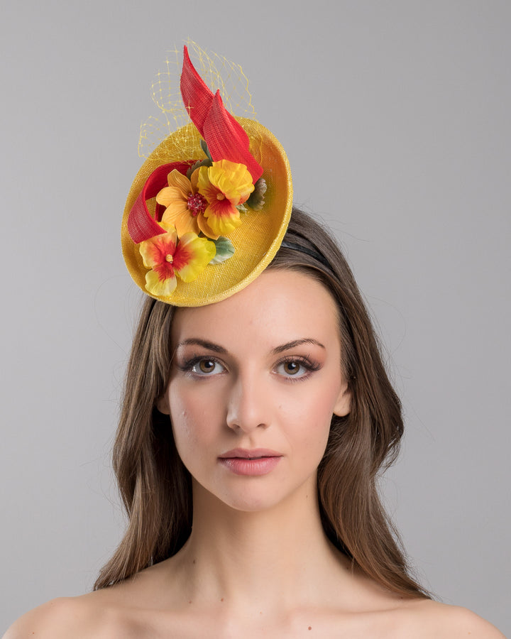 The model is showcasing the front side close up of the yellow sinamay mini fascinator with a shallow oval base, decorated with vibrant yellow and orange flowers, sculpted red sinamay loops, and a delicate birdcage veil, mounted on a slim headband. Designed and made by Merve Bayindir Millinery.

