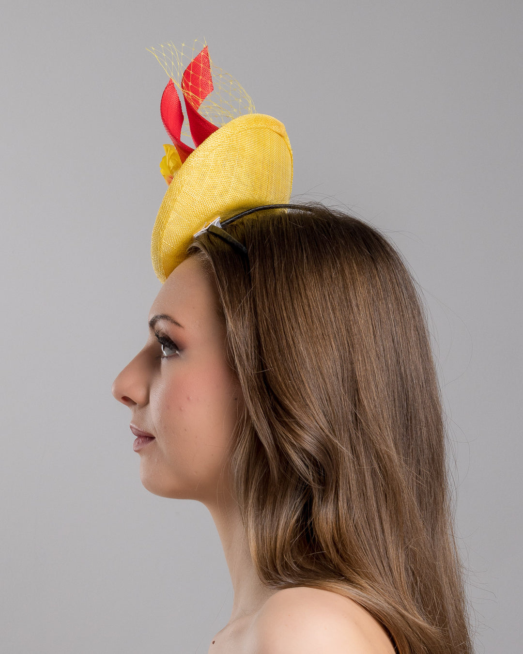 The model is showcasing the left side close up of the yellow sinamay mini fascinator with a shallow oval base, decorated with vibrant yellow and orange flowers, sculpted red sinamay loops, and a delicate birdcage veil, mounted on a slim headband. Designed and made by Merve Bayindir Millinery.
