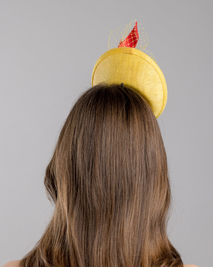 The model is showcasing the back side close up of the yellow sinamay mini fascinator with a shallow oval base, decorated with vibrant yellow and orange flowers, sculpted red sinamay loops, and a delicate birdcage veil, mounted on a slim headband. Designed and made by Merve Bayindir Millinery.
