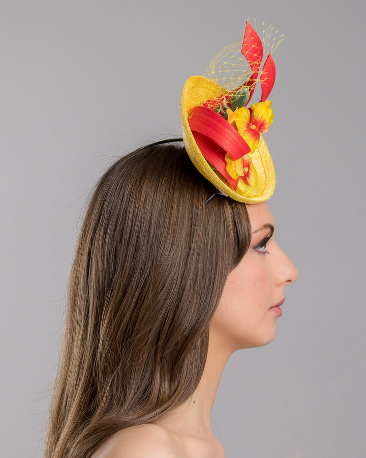 The model is showcasing the right side close up of the yellow sinamay mini fascinator with a shallow oval base, decorated with vibrant yellow and orange flowers, sculpted red sinamay loops, and a delicate birdcage veil, mounted on a slim headband. Designed and made by Merve Bayindir Millinery.
