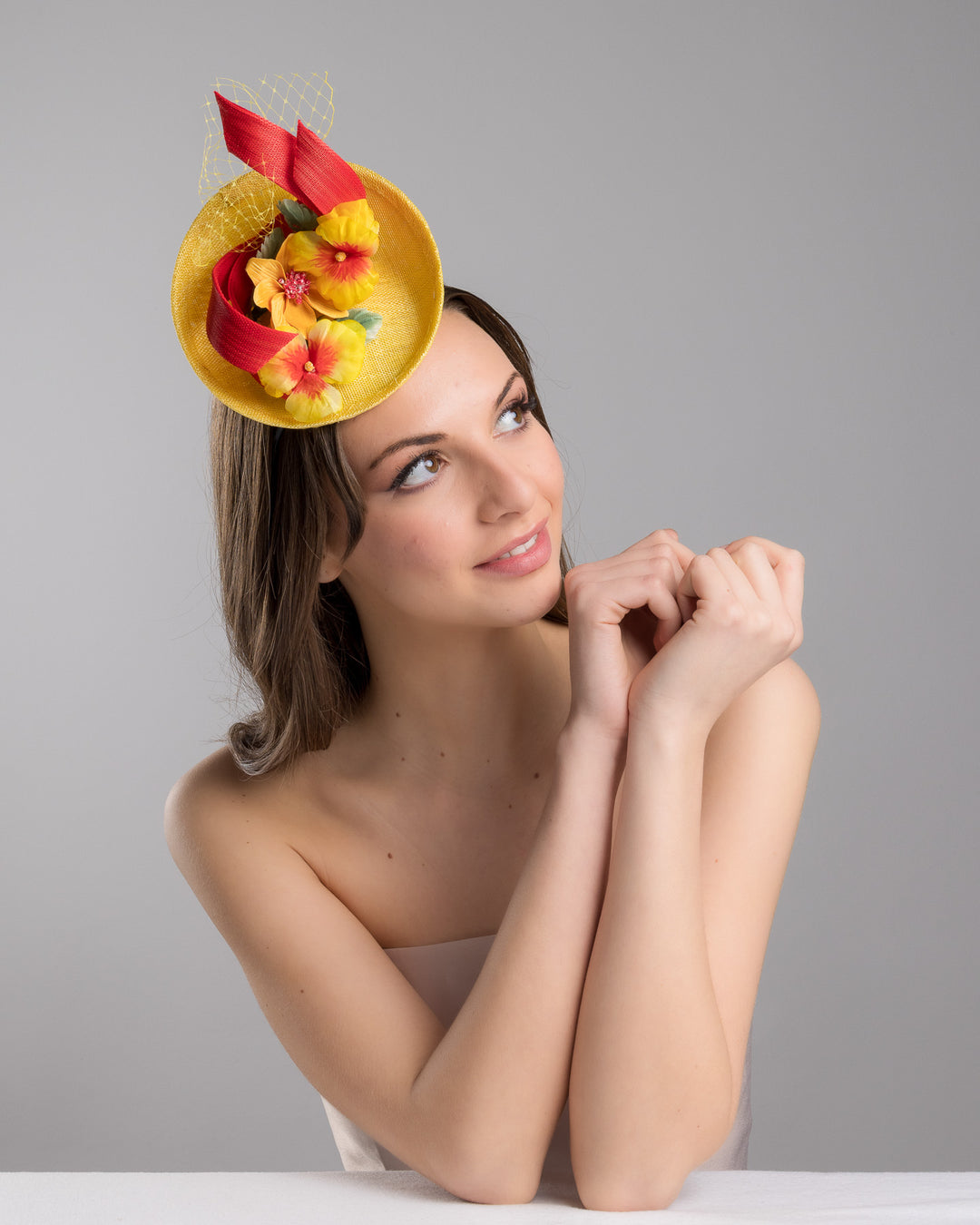 The female model is posing, wearing
Denali,  the yellow sinamay mini fascinator with a shallow oval base, decorated with vibrant yellow and orange flowers, sculpted red sinamay loops, and a delicate birdcage veil, mounted on a slim headband. Designed and made by Merve Bayindir Millinery.
