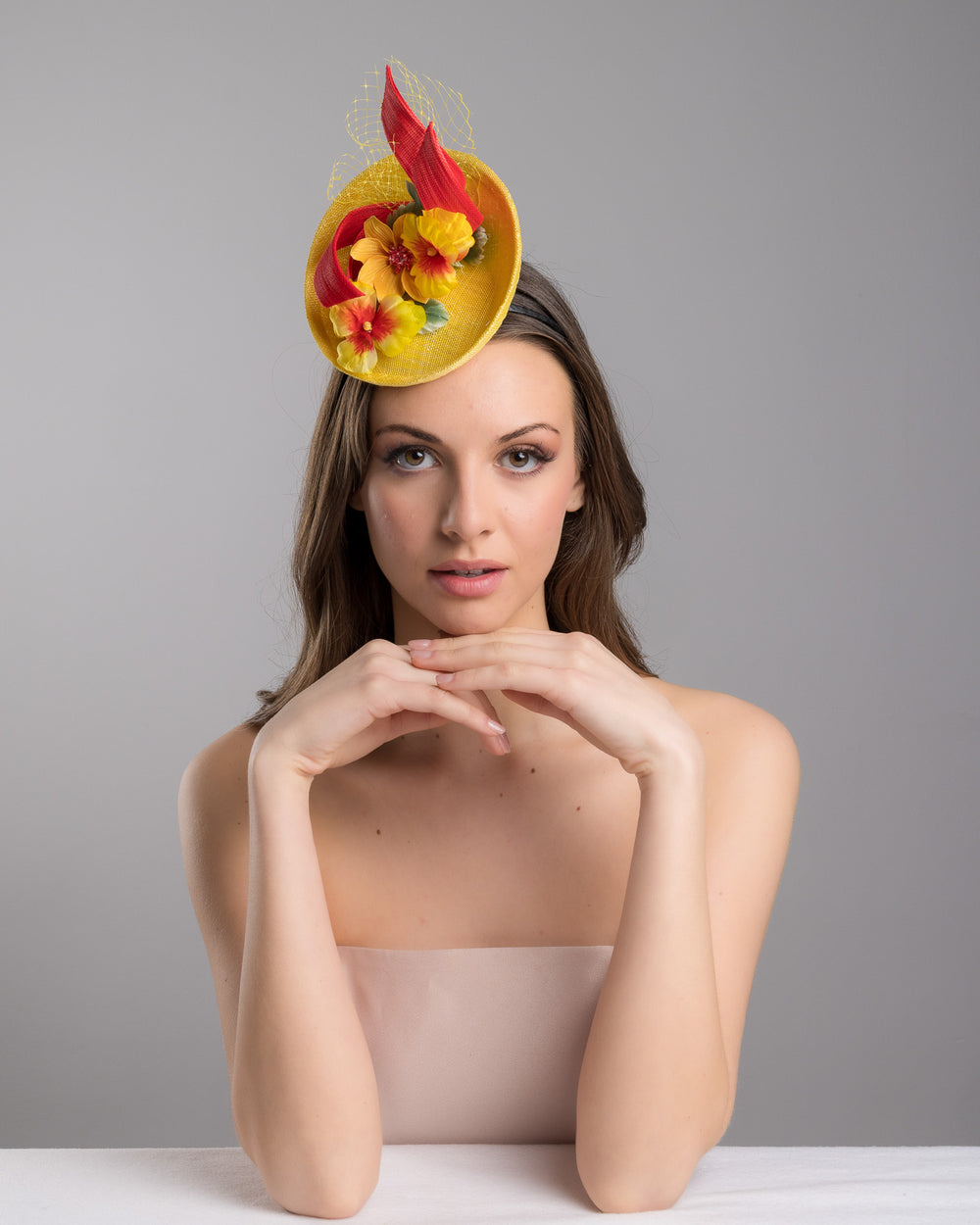 The female model is posing, wearing
Denali,  the yellow sinamay mini fascinator with a shallow oval base, decorated with vibrant yellow and orange flowers, sculpted red sinamay loops, and a delicate birdcage veil, mounted on a slim headband. Designed and made by Merve Bayindir Millinery.
