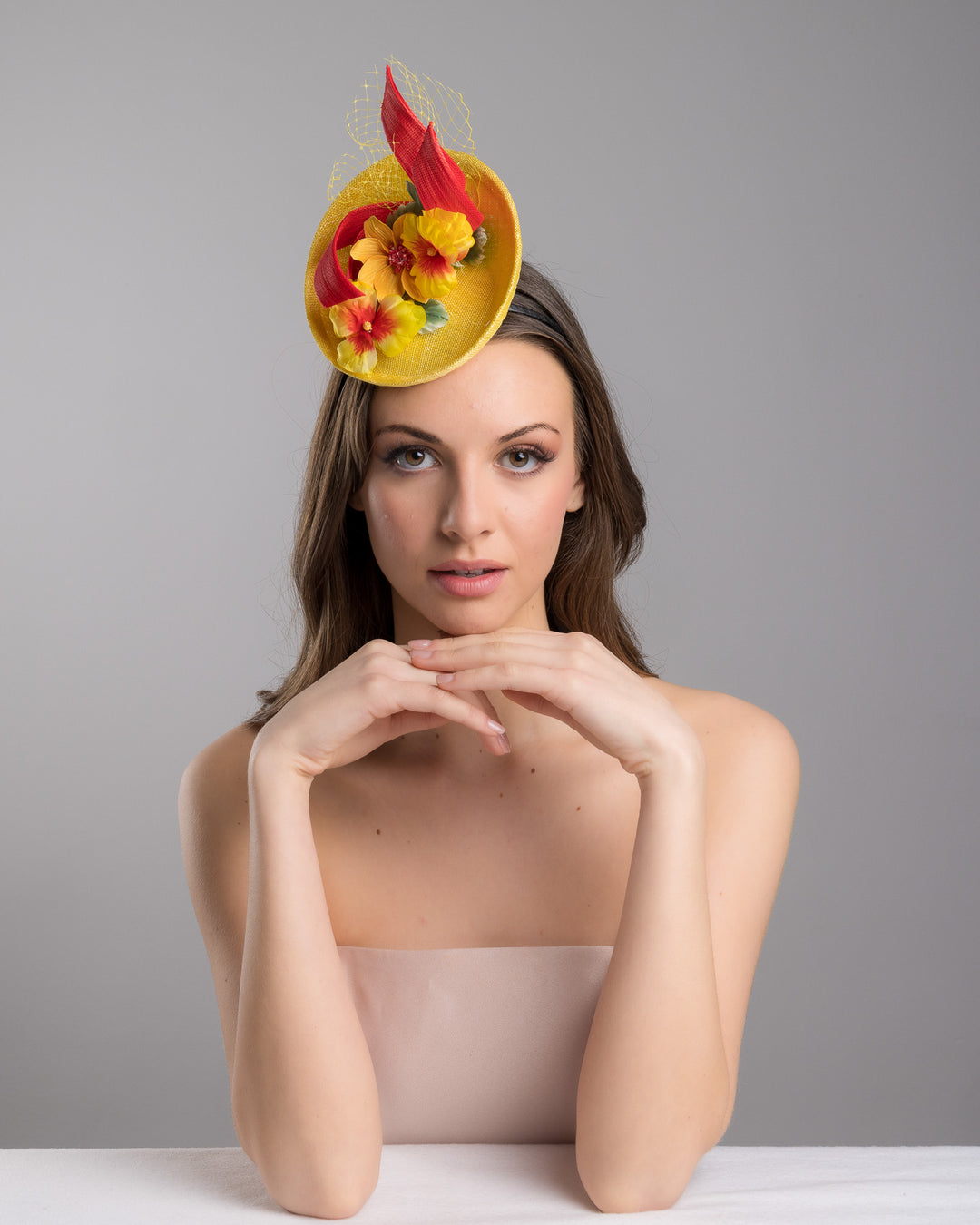 The female model is posing, wearing
Denali,  the yellow sinamay mini fascinator with a shallow oval base, decorated with vibrant yellow and orange flowers, sculpted red sinamay loops, and a delicate birdcage veil, mounted on a slim headband. Designed and made by Merve Bayindir Millinery.
