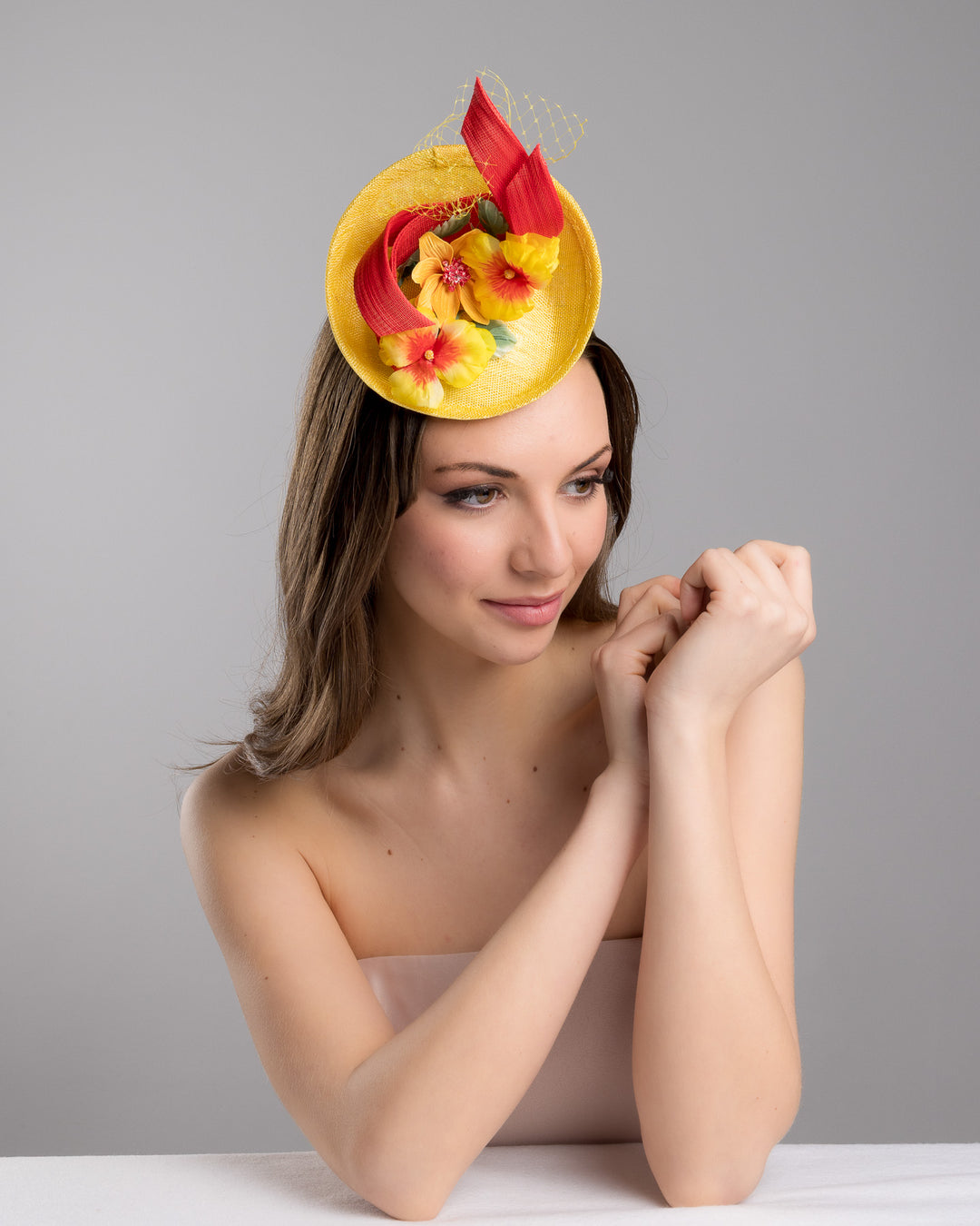 The female model is posing, wearing
Denali,  the yellow sinamay mini fascinator with a shallow oval base, decorated with vibrant yellow and orange flowers, sculpted red sinamay loops, and a delicate birdcage veil, mounted on a slim headband. Designed and made by Merve Bayindir Millinery.
