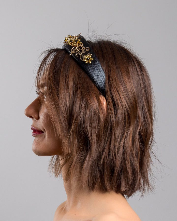 The model is showcasing the left side close up of the wide black satin headband embellished with ornate gold filigree scrollwork and small metallic floral accents, creating a regal, baroque-inspired statement design.
Designed and made by Merve Bayindir Millinery.
