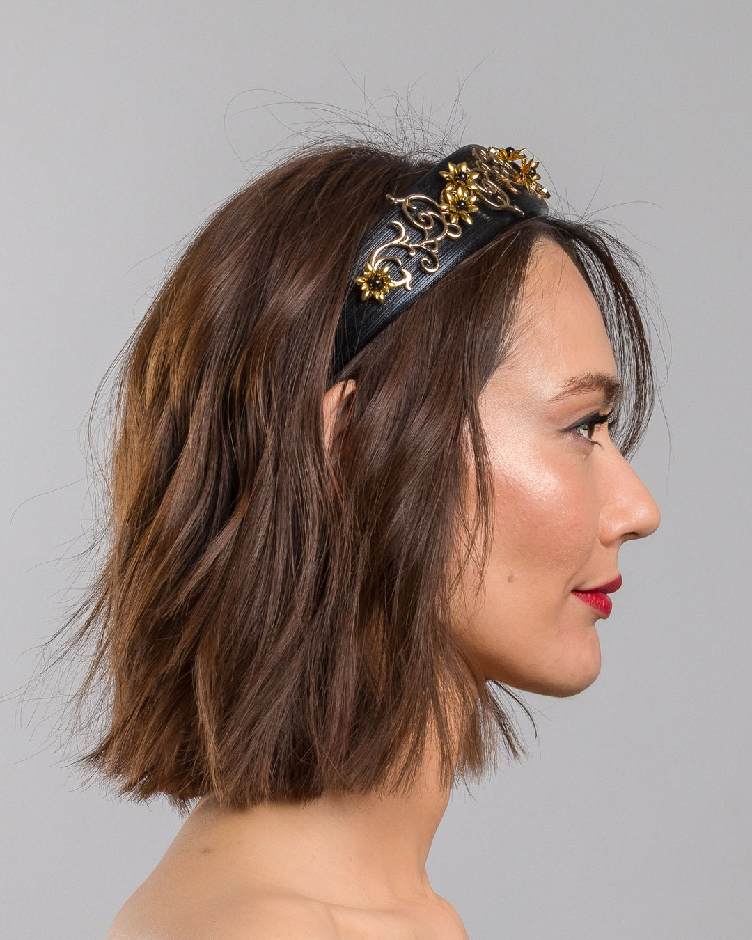 The model is showcasing the right side close up of the wide black satin headband embellished with ornate gold filigree scrollwork and small metallic floral accents, creating a regal, baroque-inspired statement design.
Designed and made by Merve Bayindir Millinery.
