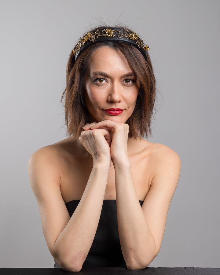 The female model is posing, wearing Doreal, the wide black satin headband embellished with ornate gold filigree scrollwork and small metallic floral accents, creating a regal, baroque-inspired statement design.
Designed and made by Merve Bayindir Millinery.