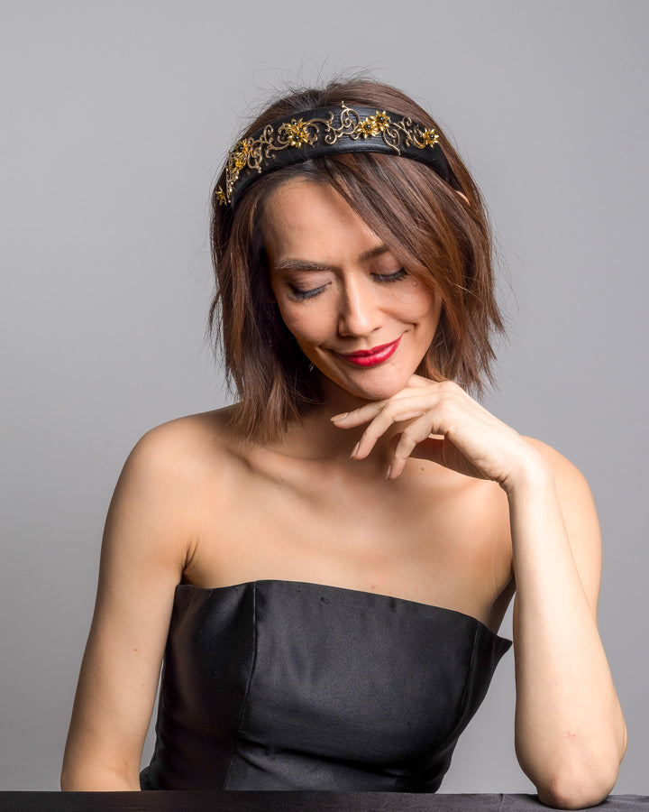The female model is posing, wearing Doreal, the wide black satin headband embellished with ornate gold filigree scrollwork and small metallic floral accents, creating a regal, baroque-inspired statement design.
Designed and made by Merve Bayindir Millinery.
