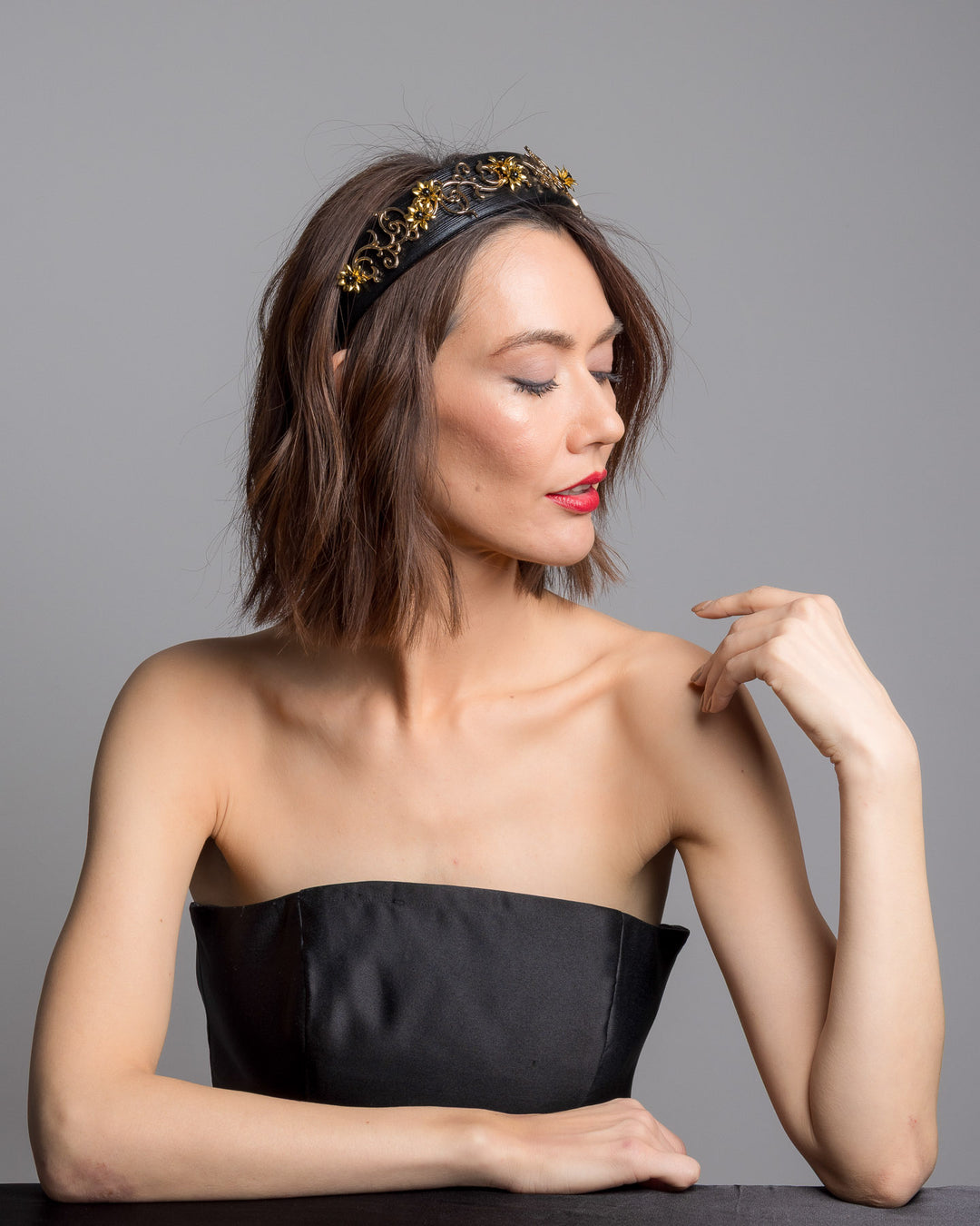 The female model is posing, wearing Doreal, the wide black satin headband embellished with ornate gold filigree scrollwork and small metallic floral accents, creating a regal, baroque-inspired statement design.
Designed and made by Merve Bayindir Millinery.