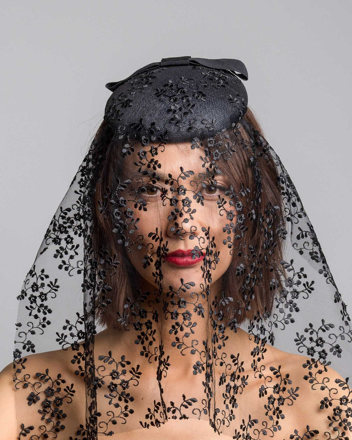 The model is showcasing the front side close up of the black round hat with a structured crown and subtle floral texture, topped with a wide ribbon band and finished with a long sheer lace veil embroidered with delicate black floral patterns. Designed and made by Merve Bayindir Millinery.