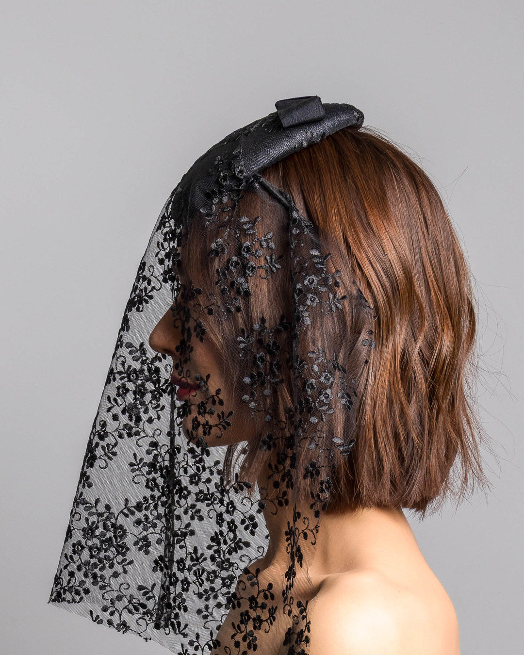 The model is showcasing the left side close up of the black round hat with a structured crown and subtle floral texture, topped with a wide ribbon band and finished with a long sheer lace veil embroidered with delicate black floral patterns. Designed and made by Merve Bayindir Millinery.