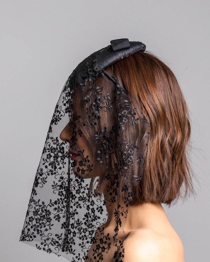 The model is showcasing the left side close up of the black round hat with a structured crown and subtle floral texture, topped with a wide ribbon band and finished with a long sheer lace veil embroidered with delicate black floral patterns. Designed and made by Merve Bayindir Millinery.