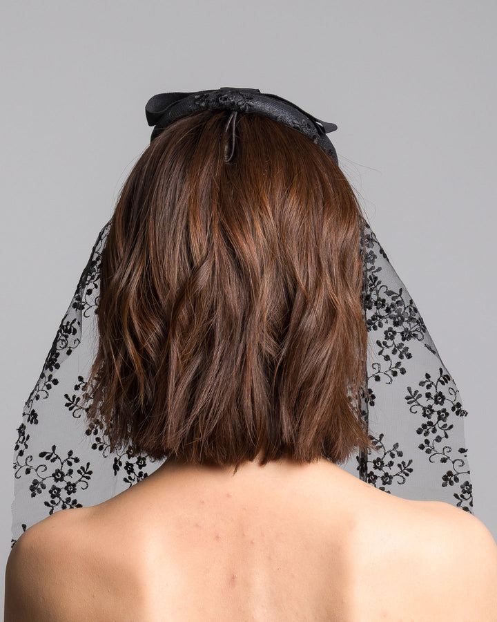 The model is showcasing the back side close up of the black round hat with a structured crown and subtle floral texture, topped with a wide ribbon band and finished with a long sheer lace veil embroidered with delicate black floral patterns. Designed and made by Merve Bayindir Millinery.