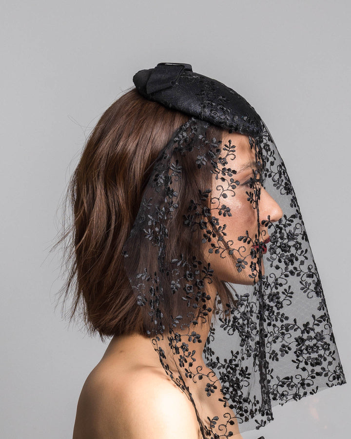 The model is showcasing the right side close up of the black round hat with a structured crown and subtle floral texture, topped with a wide ribbon band and finished with a long sheer lace veil embroidered with delicate black floral patterns. Designed and made by Merve Bayindir Millinery.