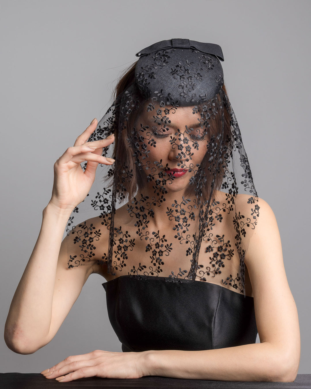 The female model is posing, wearing 
Duskier, the black round hat with a structured crown and subtle floral texture, topped with a wide ribbon band and finished with a long sheer lace veil embroidered with delicate black floral patterns. Designed and made by Merve Bayindir Millinery.