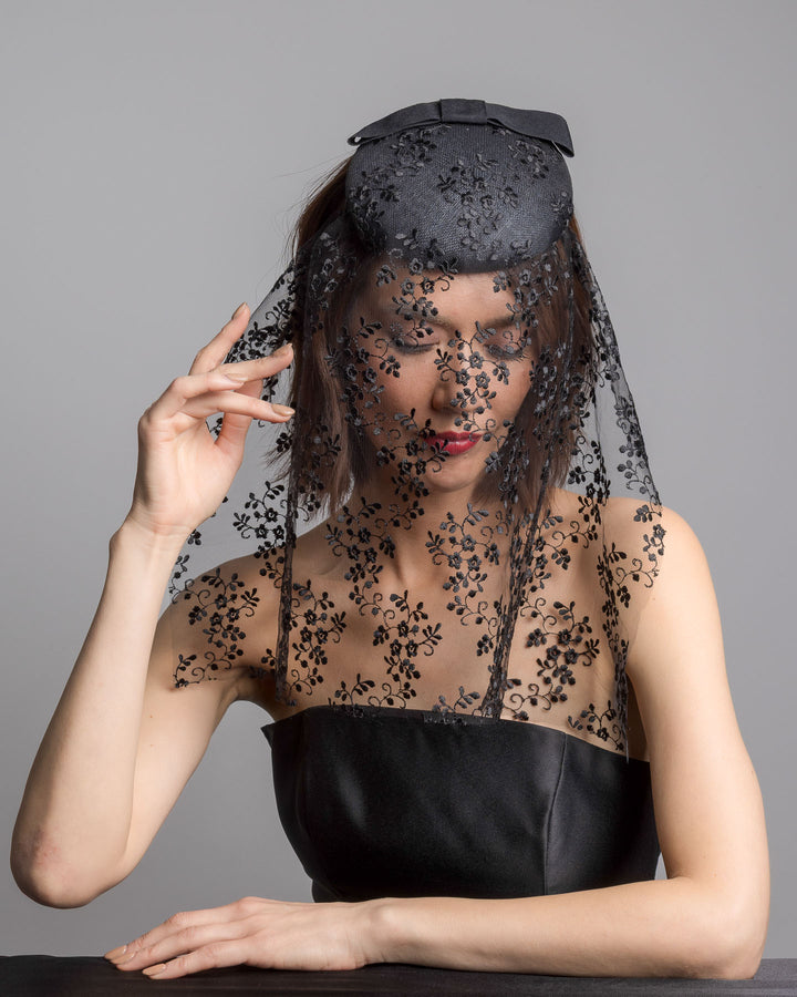 The female model is posing, wearing 
Duskier, the black round hat with a structured crown and subtle floral texture, topped with a wide ribbon band and finished with a long sheer lace veil embroidered with delicate black floral patterns. Designed and made by Merve Bayindir Millinery.