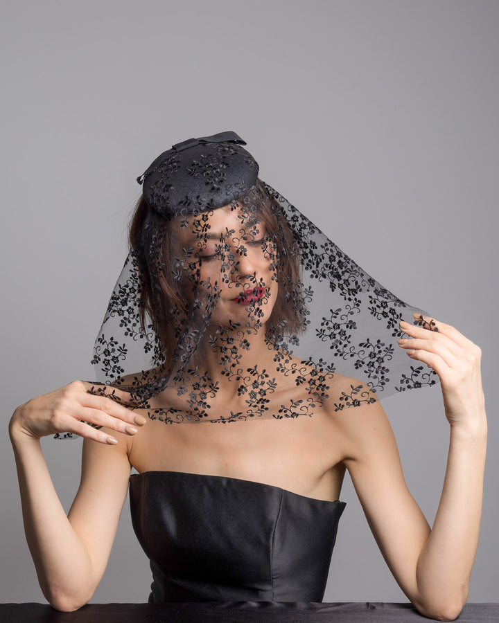 The female model is posing, wearing 
Duskier, the black round hat with a structured crown and subtle floral texture, topped with a wide ribbon band and finished with a long sheer lace veil embroidered with delicate black floral patterns. Designed and made by Merve Bayindir Millinery.