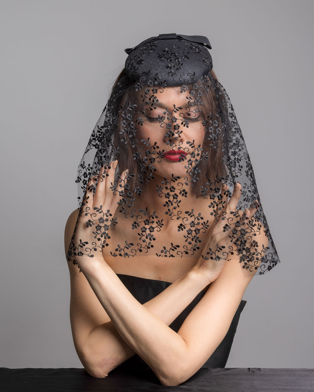 The female model is posing, wearing 
Duskier, the black round hat with a structured crown and subtle floral texture, topped with a wide ribbon band and finished with a long sheer lace veil embroidered with delicate black floral patterns. Designed and made by Merve Bayindir Millinery.