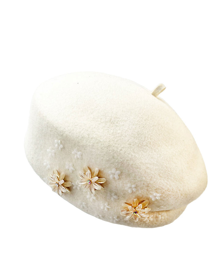 Still life picture of a white wool felt beret-style hat. This rounded design is adorned with a trio of golden, light pink floral accents, in addition to mini white flowers. Designed by Merve Bayindir.