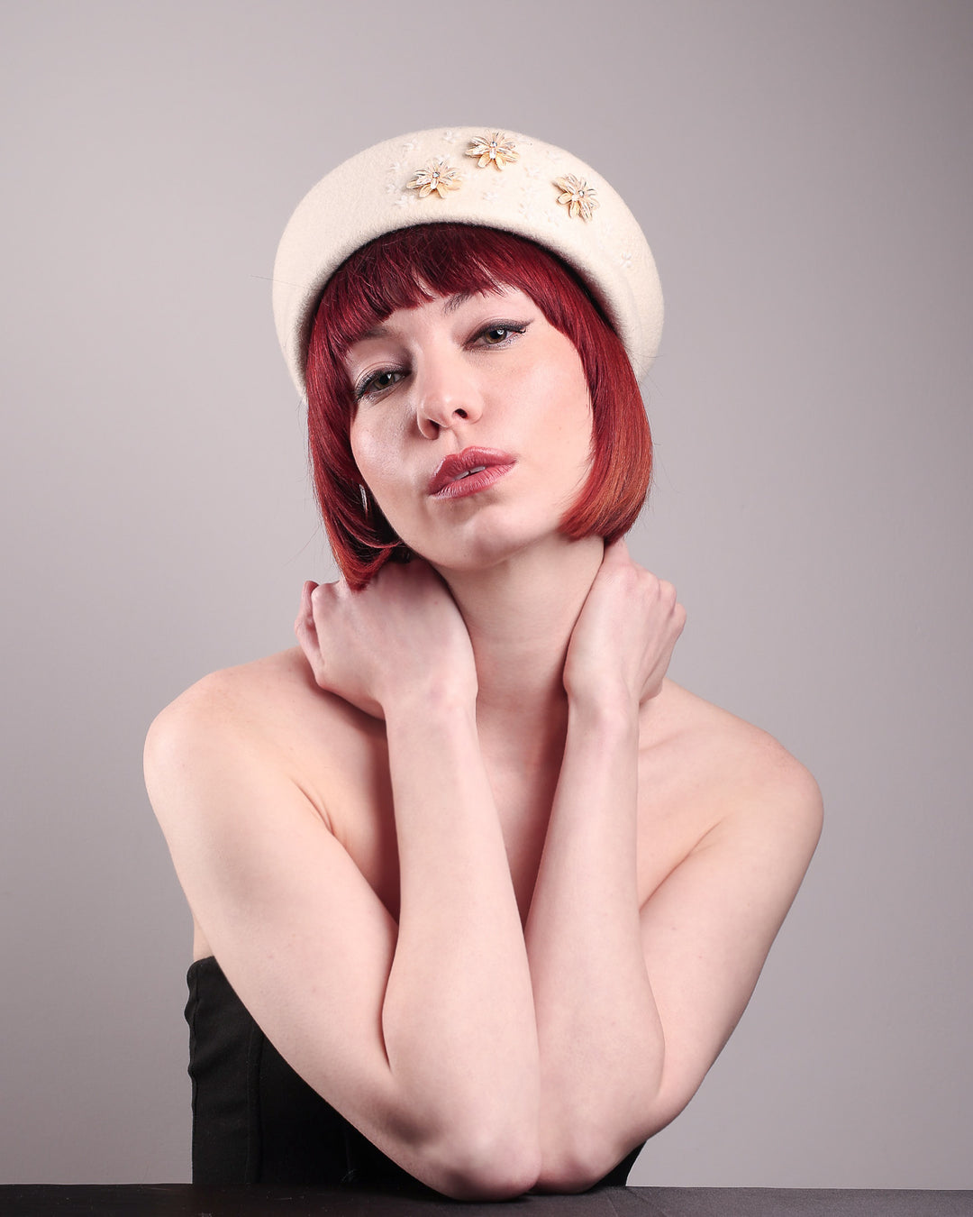 The female model is wearing a white wool felt beret-style hat, showcasing the front below angle of the hat. This rounded design is adorned with a trio of golden, light pink floral accents, in addition to mini white flowers. Designed by Merve Bayindir.