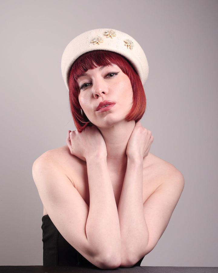 The female model is wearing a white wool felt beret-style hat, showcasing the front below angle of the hat. This rounded design is adorned with a trio of golden, light pink floral accents, in addition to mini white flowers. Designed by Merve Bayindir.