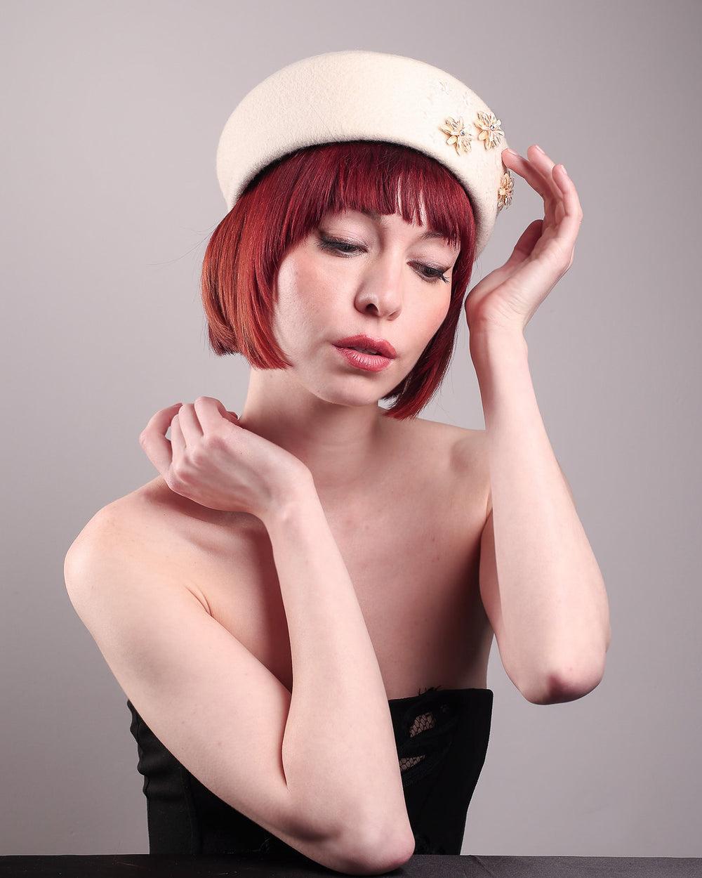 The female model is wearing a white wool felt beret-style hat, showcasing the front right view of the hat. This rounded design is adorned with a trio of golden, light pink floral accents, in addition to mini white flowers. Designed by Merve Bayindir.