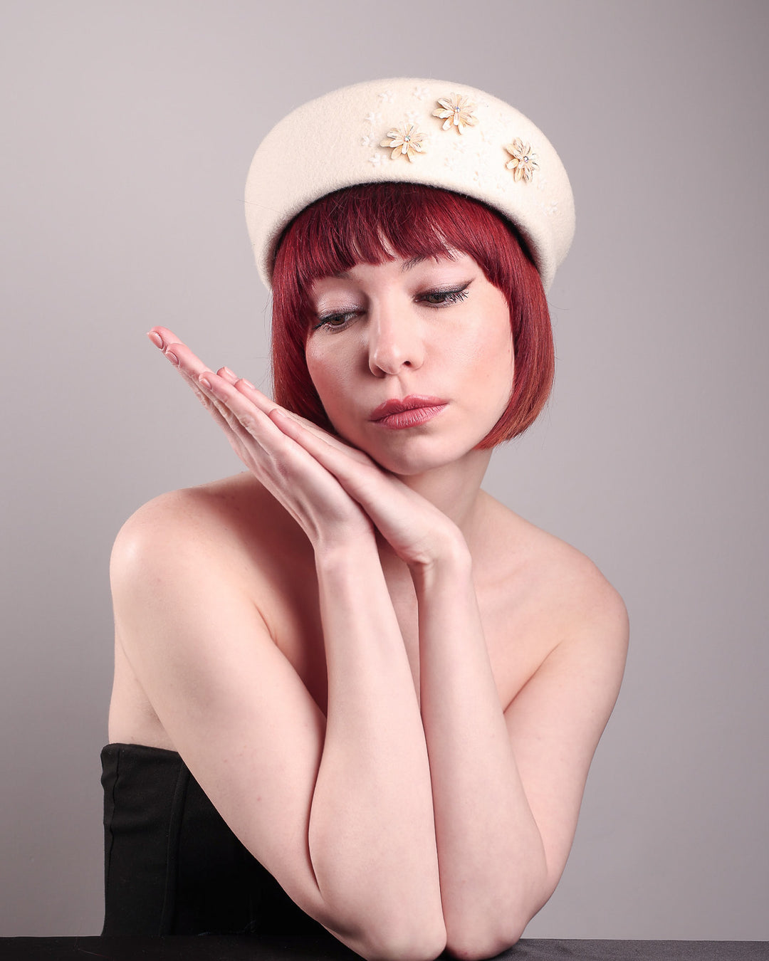 The female model is wearing a white wool felt beret-style hat, showcasing the front view of the hat. This rounded design is adorned with a trio of golden, light pink floral accents, in addition to mini white flowers. Designed by Merve Bayindir.