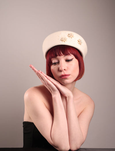 The female model is wearing a white wool felt beret-style hat, showcasing the front view of the hat. This rounded design is adorned with a trio of golden, light pink floral accents, in addition to mini white flowers. Designed by Merve Bayindir.