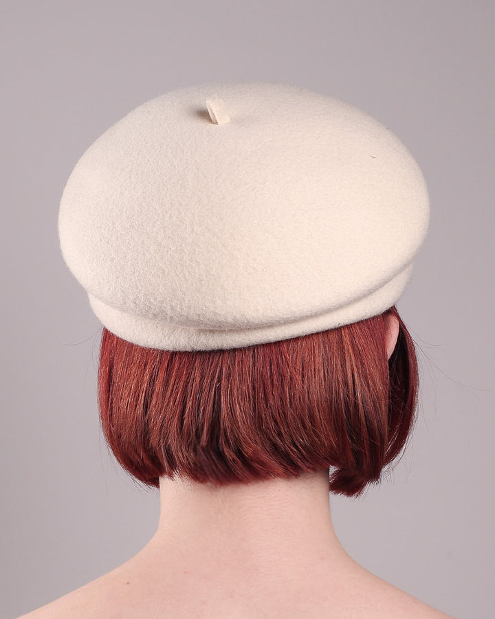 The female model is wearing a white wool felt beret-style hat, showcasing the back of the hat. This rounded design is adorned with a trio of golden, light pink floral accents, in addition to mini white flowers. Designed by Merve Bayindir.