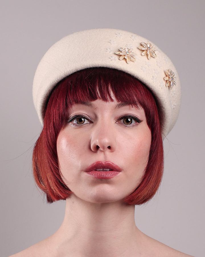 The female model is wearing a white wool felt beret-style hat, showcasing the front close up of the hat. This rounded design is adorned with a trio of golden, light pink floral accents, in addition to mini white flowers. Designed by Merve Bayindir.