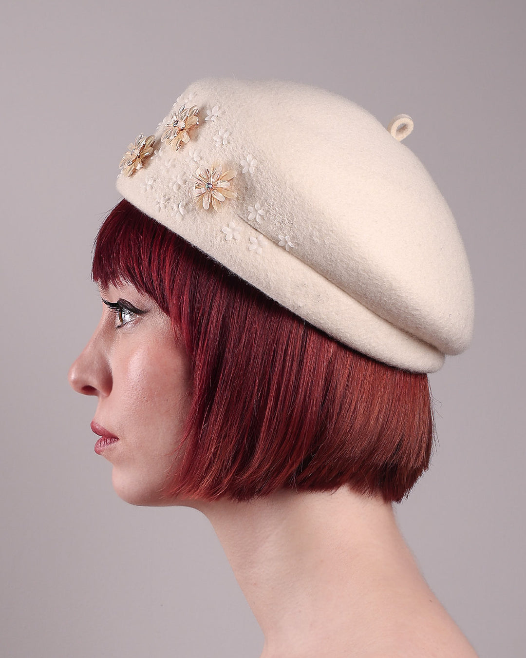 The female model is wearing a white wool felt beret-style hat, showcasing the left side close up of the hat. This rounded design is adorned with a trio of golden, light pink floral accents, in addition to mini white flowers. Designed by Merve Bayindir.