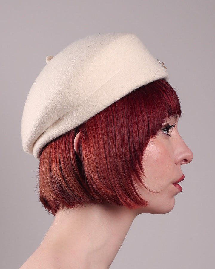 The female model is wearing a white wool felt beret-style hat, showcasing the right side close up of the hat. This rounded design is adorned with a trio of golden, light pink floral accents, in addition to mini white flowers. Designed by Merve Bayindir.