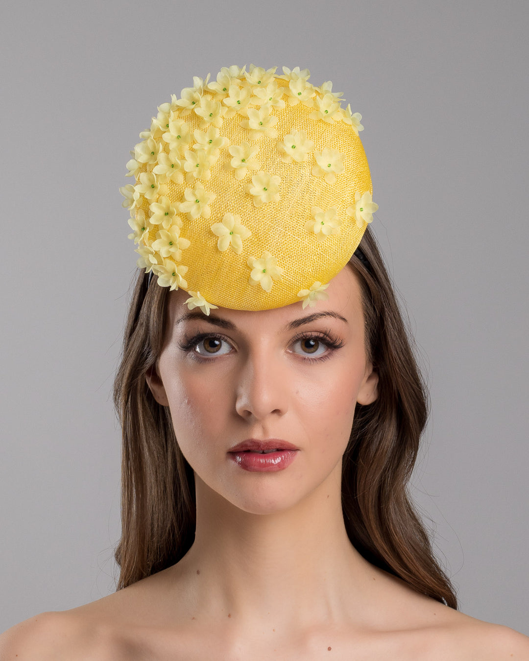 Model is showcasing the front side close up of yellow button shaped fascinator, decorated with yellow organza flowers and green beads. Designed and made by Merve Bayindir Millinery.