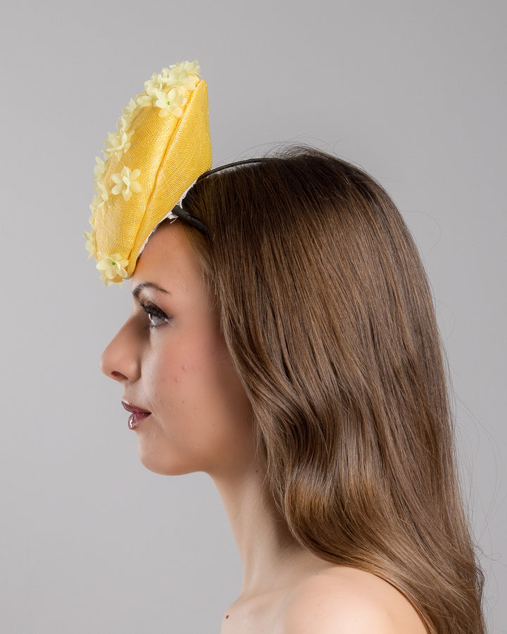 Model is showcasing the left side close up of yellow button shaped fascinator, decorated with yellow organza flowers and green beads. Designed and made by Merve Bayindir Millinery.
