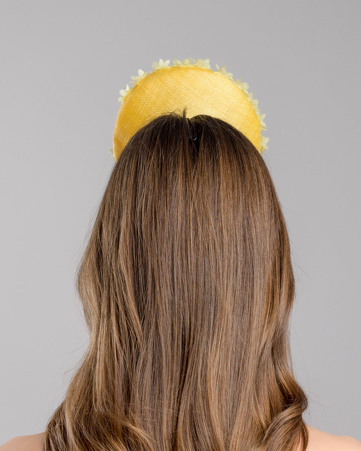 Model is showcasing the back side close up of yellow button shaped fascinator, decorated with yellow organza flowers and green beads. Designed and made by Merve Bayindir Millinery.