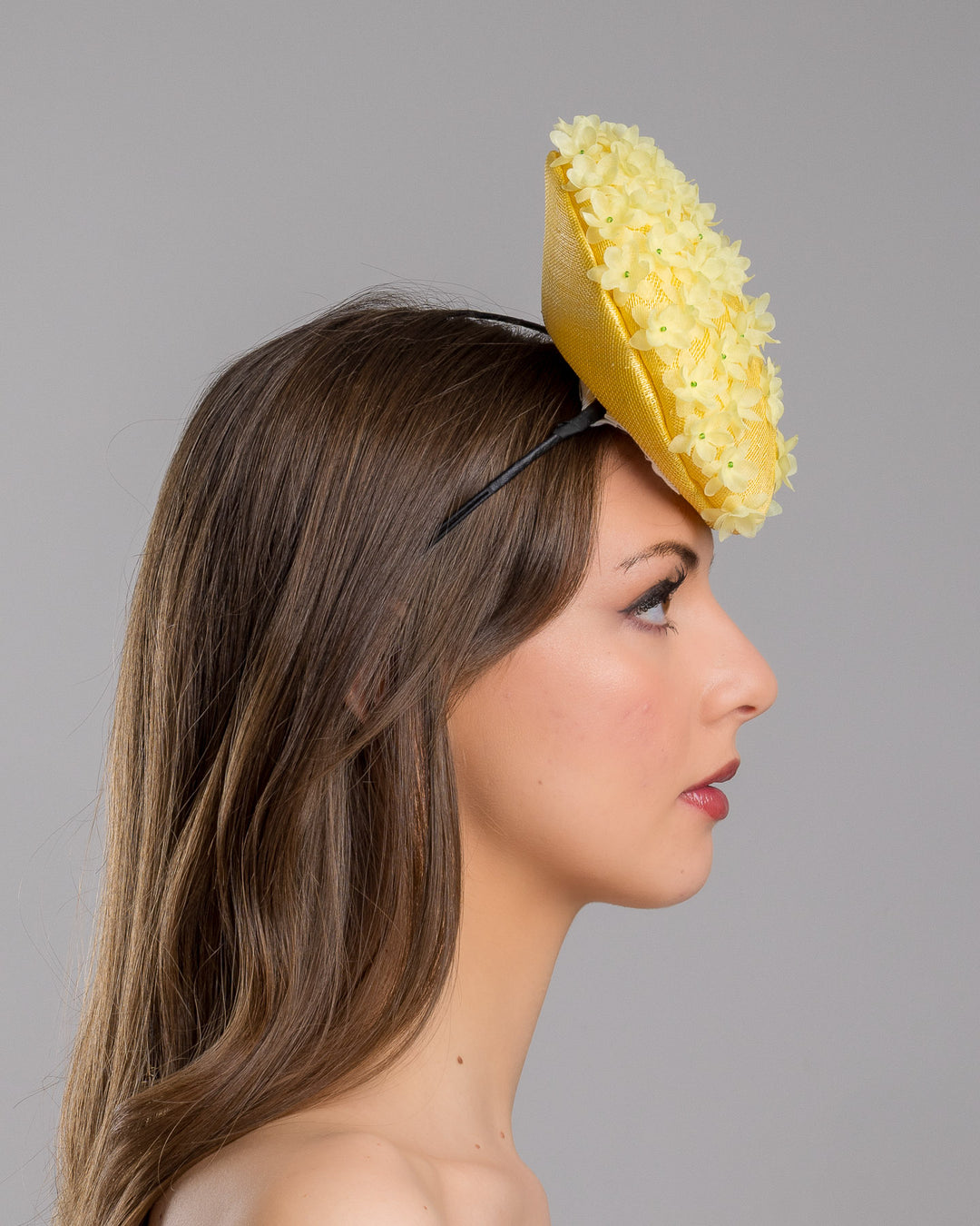 Model is showcasing the right side close up of yellow button shaped fascinator, decorated with yellow organza flowers and green beads. Designed and made by Merve Bayindir Millinery.