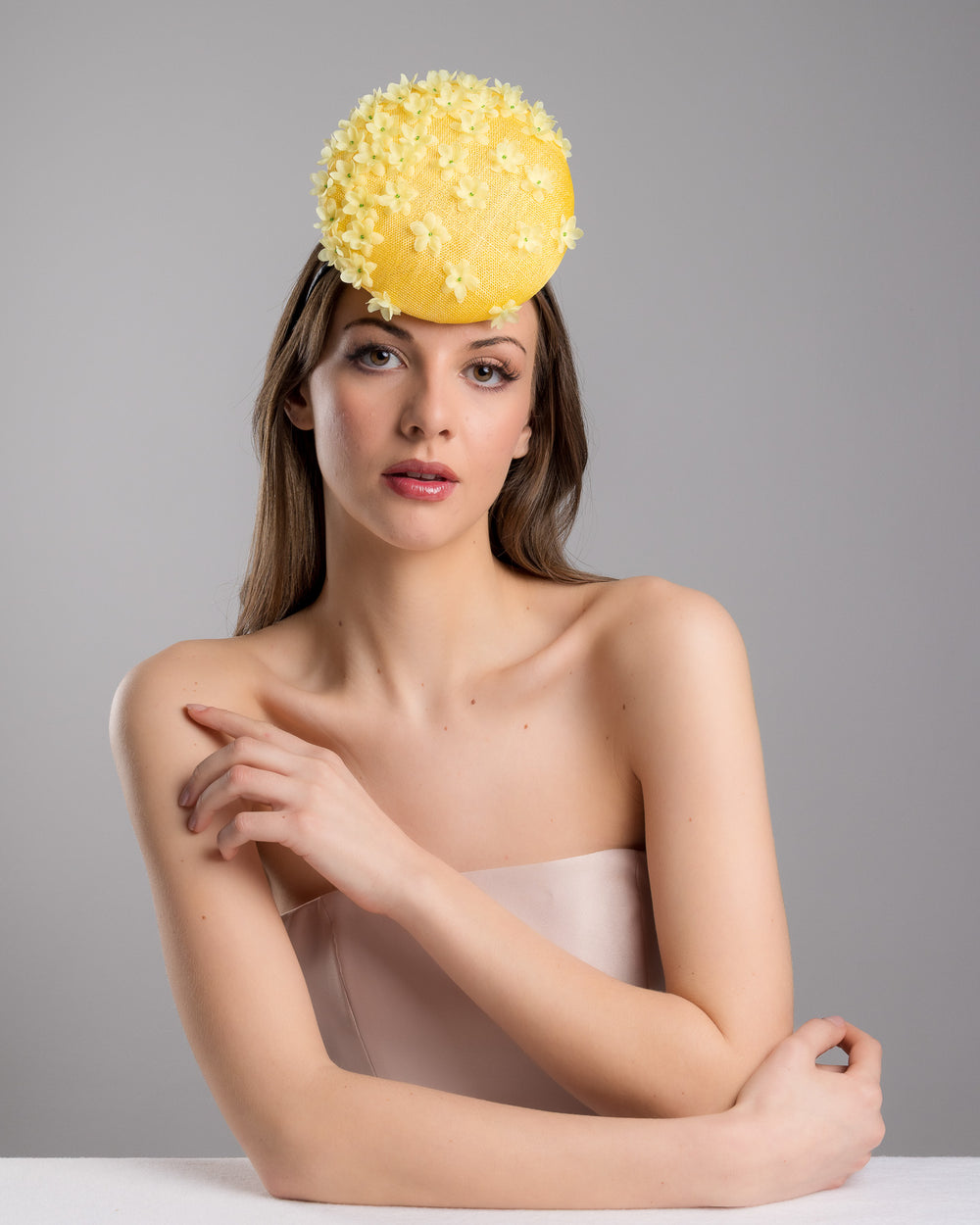 Female model is posing wearing the Dale yellow button shaped fascinator, decorated with yellow organza flowers and green beads. Designed and made by Merve Bayindir Millinery.