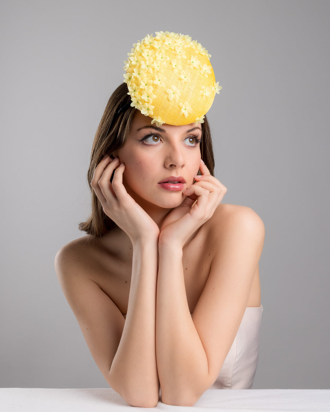 Female model is posing wearing the Dale yellow button shaped fascinator, decorated with yellow organza flowers and green beads. Designed and made by Merve Bayindir Millinery.